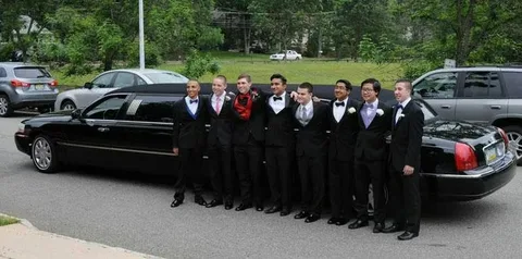 Group of young men in formal prom suits standing beside a black stretch limousine before prom night transportation.